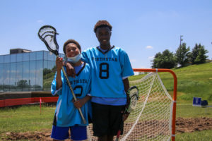 in the picture a shorter light skin black boy smiles holding a lacrosse stick, a taller dark skin black boy smiles with his arm around his shoulder. they are outside with green grass and blue skies with a building a lacrosse net in the background