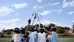 group of young men in circle holding up lacrosse sticks on outdoor field with blue skies