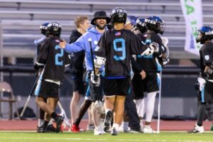 A happy coach smiles before exchanging a high five with a uniformed player on the lacrosse field